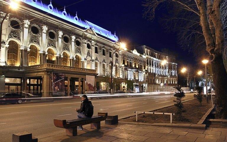 A man sits on a bench in front of a building at night in Tbilisi, Georgia.