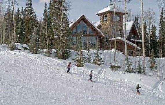 A group of people skiing down a snowy slope in front of a log cabin at Alpen Ridge Villa.