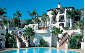 Bacara Resort & Spa welcomes you with open arms to a swimming pool in front of a white building surrounded by palm trees.