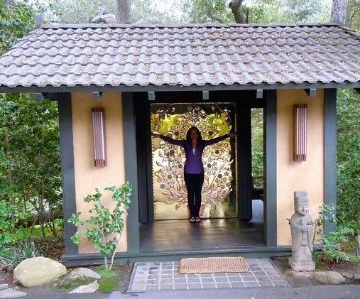 A woman standing in front of a wooden hut in the woods. Keywords: Golden Door, heaven on earth.